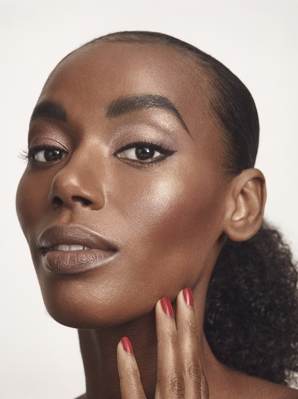 Close-up portrait of a Black female adult with neutral expression, red nails, and glossy makeup against a light neutral backdrop.