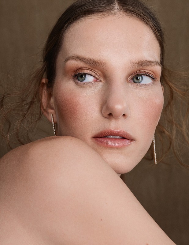 Close-up portrait of a fair-skinned adult female with neutral expression, light makeup, delicate earrings, and tousled hair against a tan backdrop.