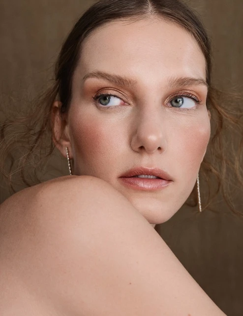 Close-up portrait of a fair-skinned adult female with neutral expression, light makeup, delicate earrings, and tousled hair against a tan backdrop.