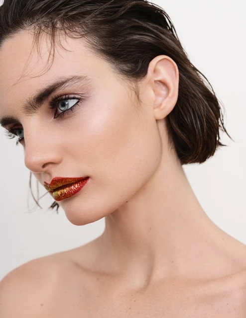 Close-up profile of a young adult female with red and gold glitter lips, short dark hair, bare shoulders, against a neutral background.