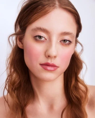 Close-up portrait of a young woman with wavy auburn hair, flushed pink cheeks, and natural makeup against a plain white background.