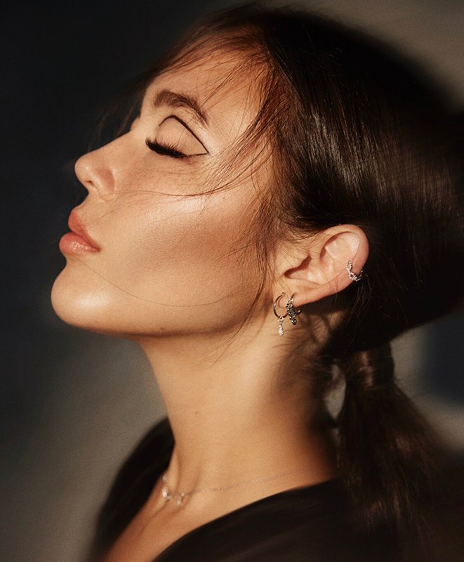 Close-up profile of a woman with closed eyes, graphic eyeliner, and styled hair featuring delicate earrings by hairstylist Destiny Venice.