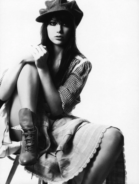 Black and white portrait of a woman with dark hair wearing a cap, polka dot shirt, and boots, sitting on a chair in a photo studio.