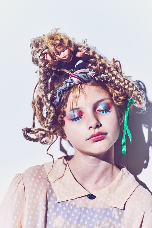 Portrait of a girl with elaborate blue and red eye makeup, dolls in her hair, wearing a beige polka dot blouse, against a white background.