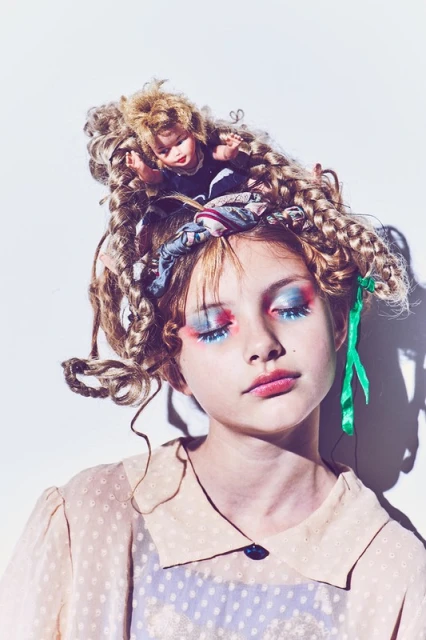 Portrait of a girl with elaborate blue and red eye makeup, dolls in her hair, wearing a beige polka dot blouse, against a white background.
