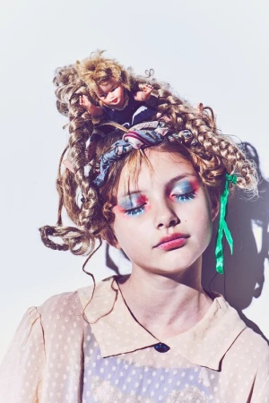 Portrait of a girl with elaborate blue and red eye makeup, dolls in her hair, wearing a beige polka dot blouse, against a white background.