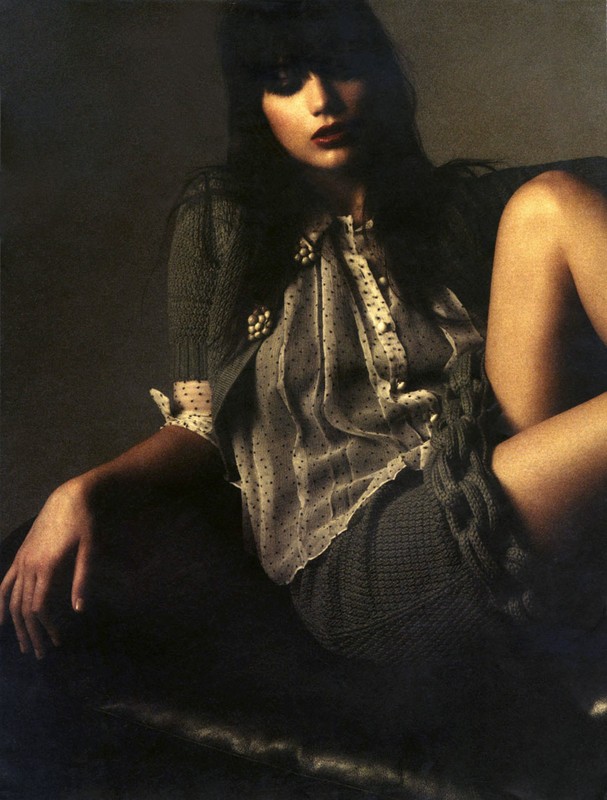 A brunette woman with dark makeup poses on a dark couch wearing a polka dot blouse and knitted sweater and skirt.