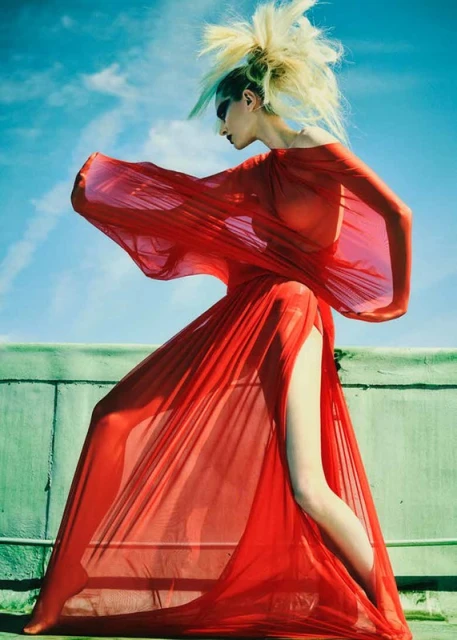 Woman in a flowing red dress poses on a rooftop with a blue sky. The dress is sheer, revealing her silhouette. Fashion shot.