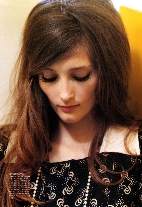 Portrait of a brunette female model with long hair, wearing a patterned top with a white collar and pearl necklace, looking downward.