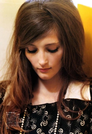 Portrait of a brunette female model with long hair, wearing a patterned top with a white collar and pearl necklace, looking downward.