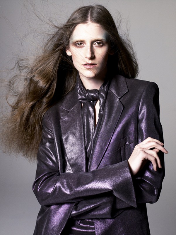 Portrait of a fair-skinned adult woman with long brown hair, wearing a purple, shiny blazer and tie, gazing intently at the camera in a studio shot.