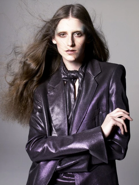 Portrait of a fair-skinned adult woman with long brown hair, wearing a purple, shiny blazer and tie, gazing intently at the camera in a studio shot.