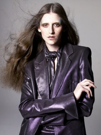 Portrait of a fair-skinned adult woman with long brown hair, wearing a purple, shiny blazer and tie, gazing intently at the camera in a studio shot.