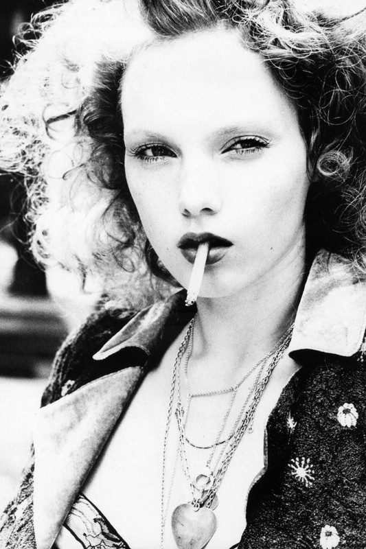Black and white portrait of a curly-haired woman smoking a cigarette, wearing a patterned jacket and layered necklaces with a heart pendant.