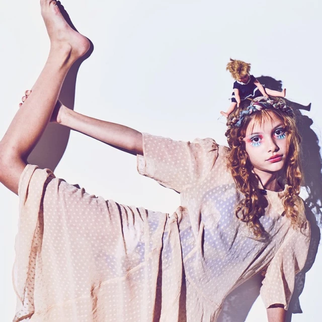A young person in a light-peach dress poses against a white background with leg raised. A doll sits on their head.
