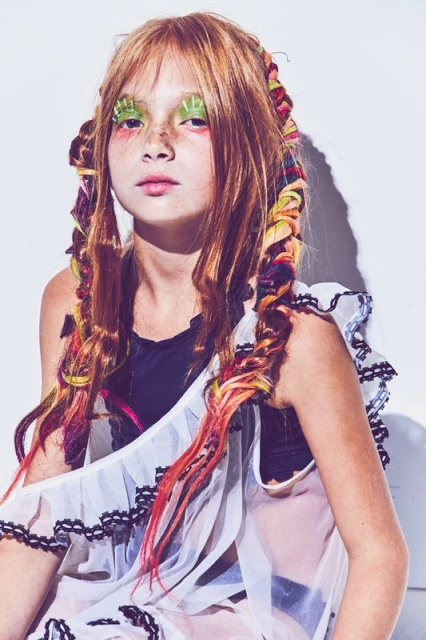 Young girl with freckles and braids wearing makeup and a frilly dress poses in a studio against a white backdrop.