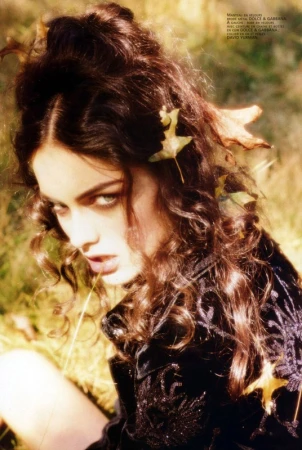 Portrait of a young woman with wavy brown hair and leaves in her hair, wearing a black embroidered jacket outdoors.
