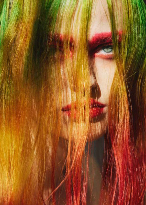 Close-up portrait of a woman with vibrant multicolored hair and red makeup, partially obscuring her face in a beauty style.