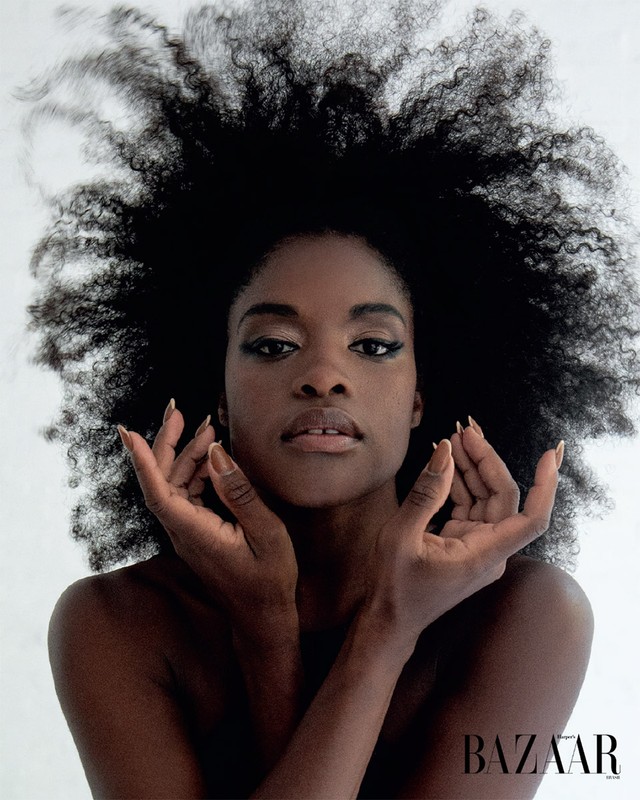 Striking portrait of a Black woman with voluminous afro hair, makeup, and manicured nails, set against a clean white background in a beauty shot.