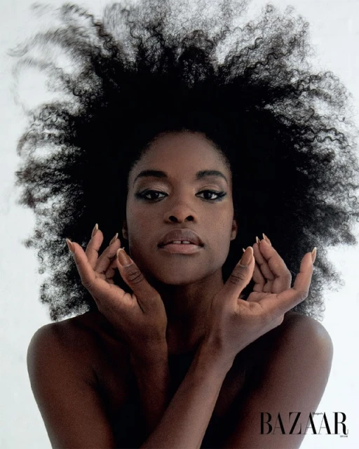 Striking portrait of a Black woman with voluminous afro hair, makeup, and manicured nails, set against a clean white background in a beauty shot.