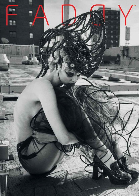 Monochrome image of a model with a futuristic hairstyle, wearing a dark, semi-sheer outfit and platform shoes, sitting on a rooftop.