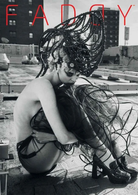 Monochrome image of a model with a futuristic hairstyle, wearing a dark, semi-sheer outfit and platform shoes, sitting on a rooftop.