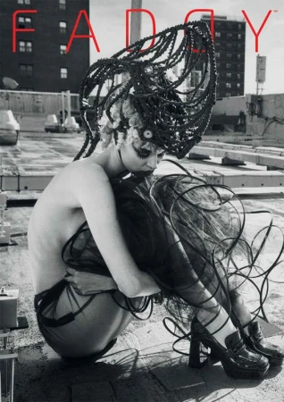 Monochrome image of a model with a futuristic hairstyle, wearing a dark, semi-sheer outfit and platform shoes, sitting on a rooftop.