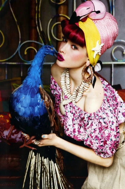 Woman in floral top with colorful headpiece holding blue peacock in a studio. Shell necklace and bold makeup complete the scene.