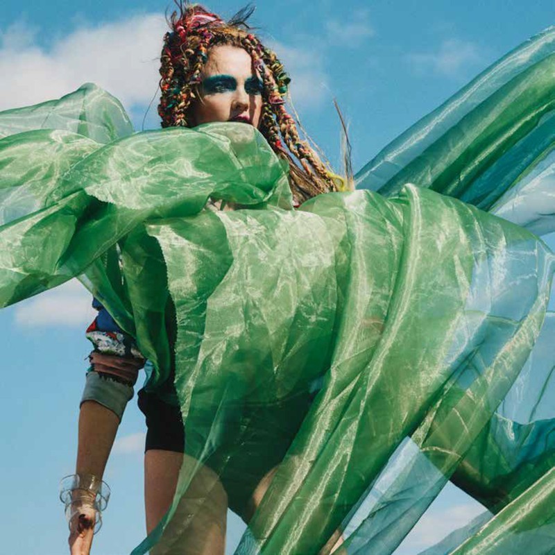 Adult female poses outdoors with colorful braids and green sheer fabric against a blue sky, showcasing makeup and a fashionable style.