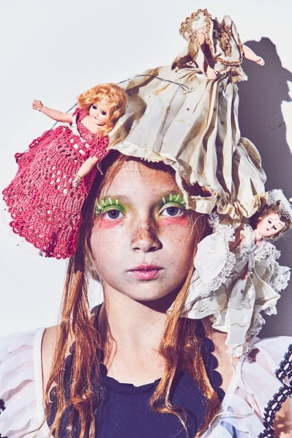 Portrait of a young person with dramatic make-up and several vintage dolls arranged as headwear, set against a white background.