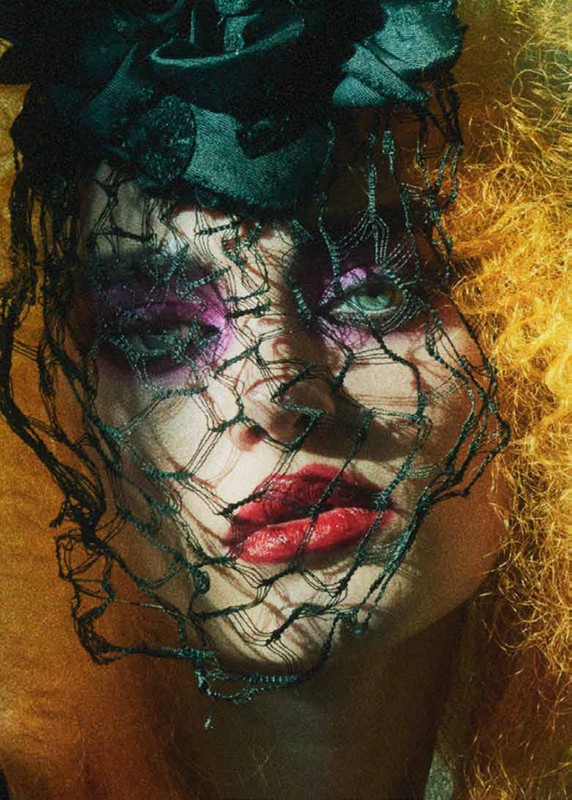 Close-up portrait of a woman with red lips, yellow hair, and dramatic purple eye makeup wearing a floral headpiece and black netting.