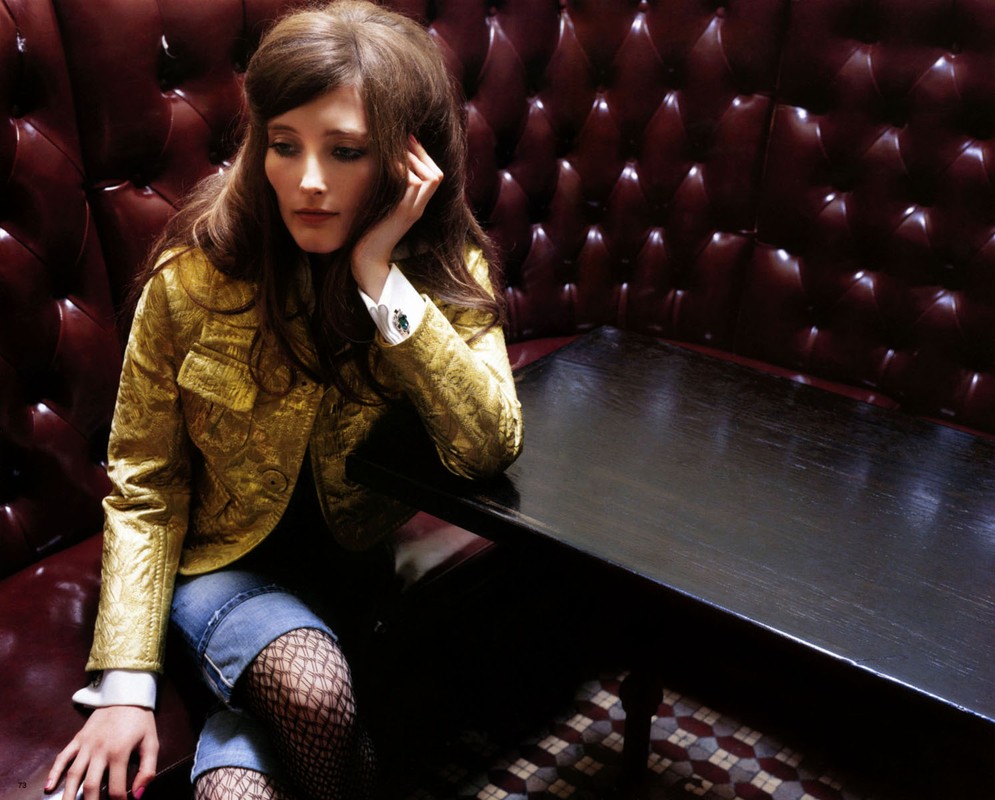 Woman with long brown hair sits in a booth wearing a gold jacket and jean shorts with black fishnet stockings in an indoor setting.