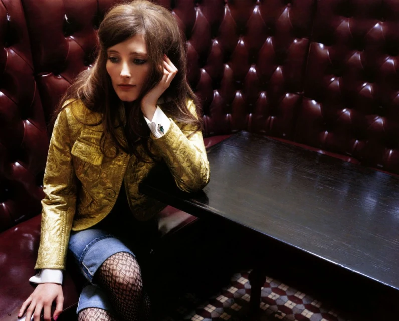 Woman with long brown hair sits in a booth wearing a gold jacket and jean shorts with black fishnet stockings in an indoor setting.