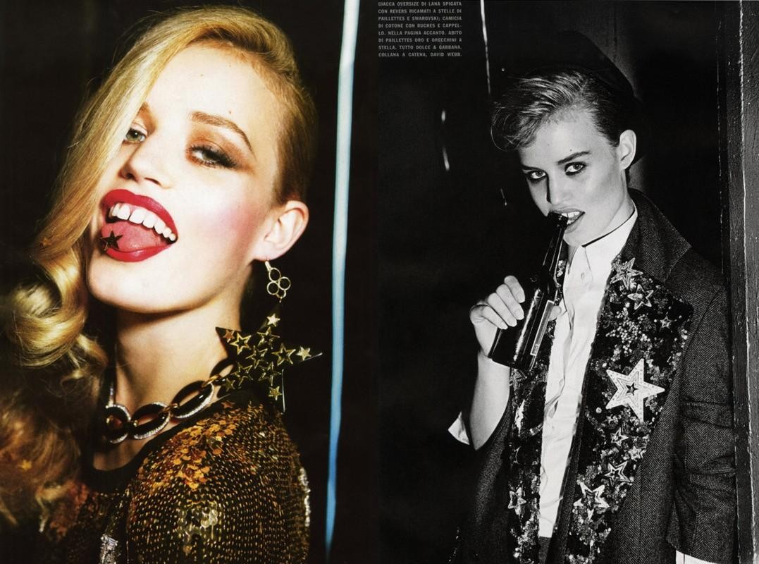 Two women pose in a studio shot with sequined clothing, statement jewelry, and a dark backdrop, one with a beer.