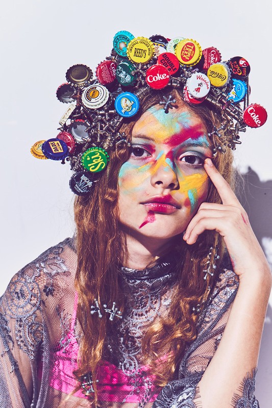 Artistic portrait of a young adult female with colorful face paint and a bottle cap crown, set against a bright, clean studio backdrop.