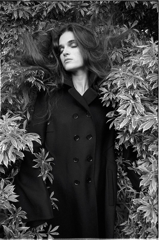 Black and white portrait of a woman in a dark coat lying among green plants, her hair spread out, eyes closed in a natural setting.