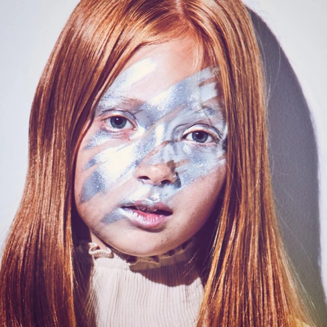 Close-up portrait of a young girl with red hair and silver face paint in a studio setting against a white background, beauty shot.