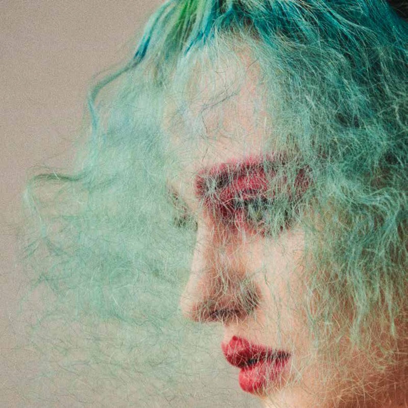 Side profile portrait of a woman with green and blue tinted hair and red lipstick, with hair partially obscuring her face.