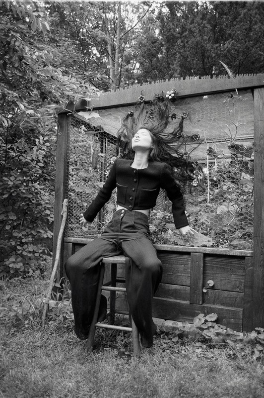 Monochrome portrait of a slender woman in a cropped jacket and wide-leg pants, seated on a stool amidst lush foliage and an arbor.