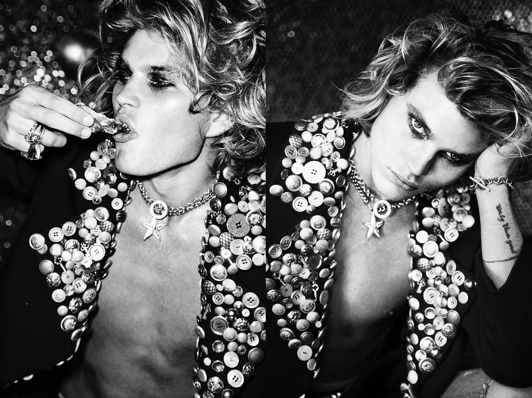 Black and white studio shot of a young adult man with blonde curly hair and a button-covered jacket in a semi-nude pose.