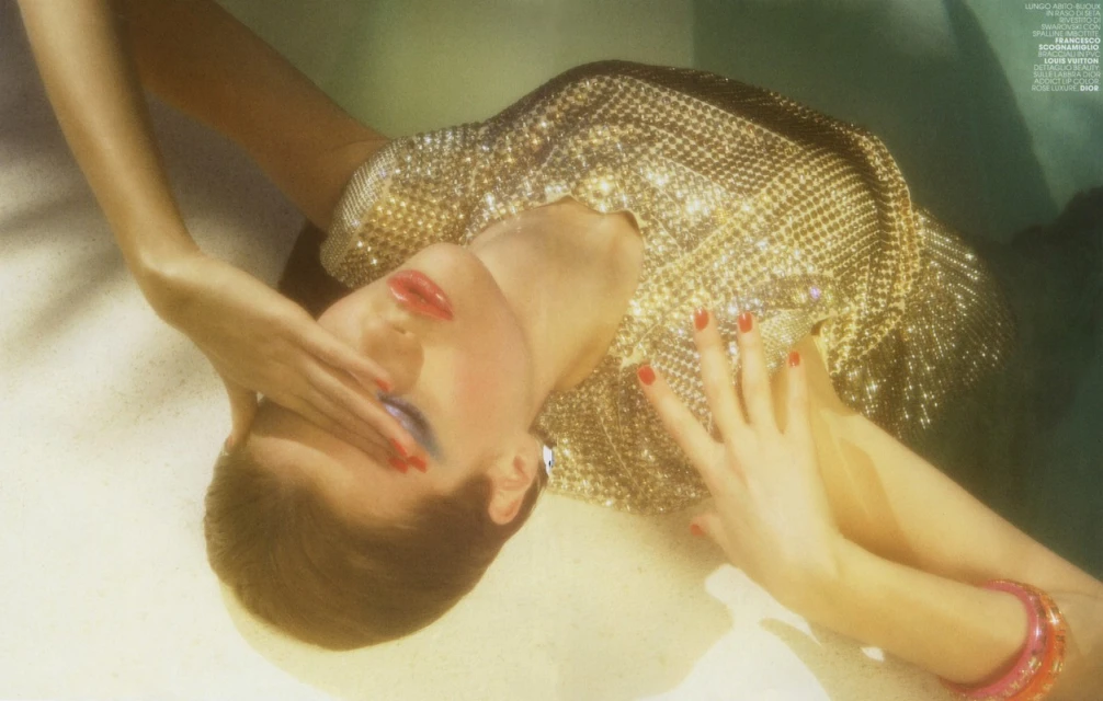 A blonde woman with blue eye makeup, red nails, and a shiny gold top poses with her hands near her face in a surreal, golden-lit scene.