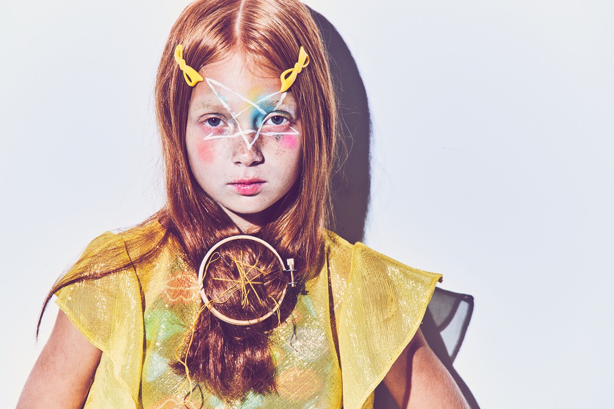 Portrait of a young girl with red hair and painted facial makeup, wearing a yellow sparkly top and posing in front of a white background.