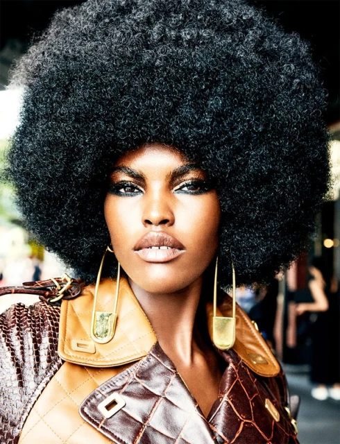 Close-up of a Black fashion model with a large afro, wearing gold safety-pin earrings and a brown leather jacket. Beauty and fashion shot.