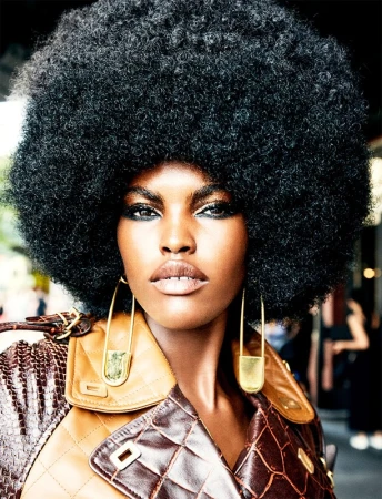 Close-up of a Black fashion model with a large afro, wearing gold safety-pin earrings and a brown leather jacket. Beauty and fashion shot.