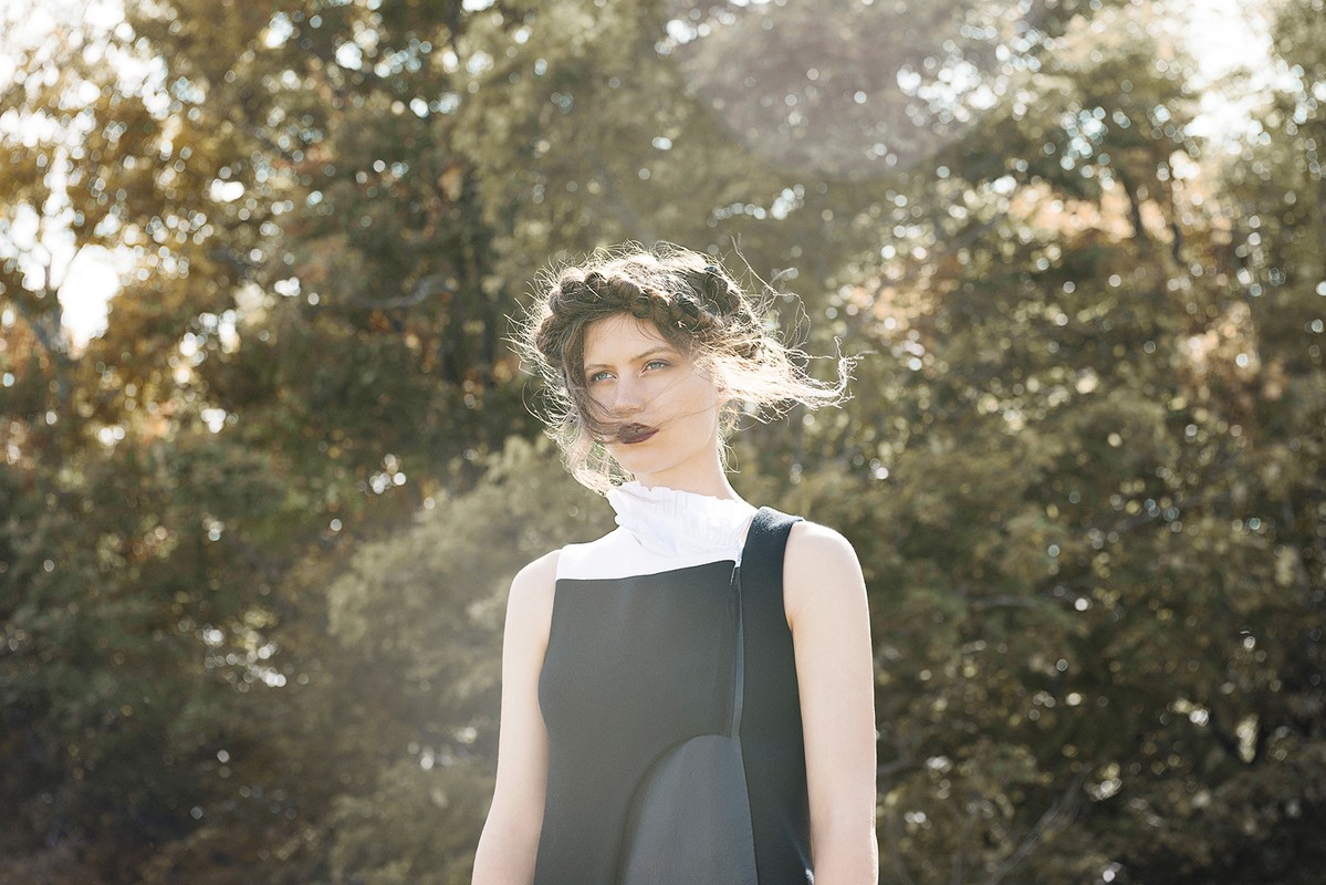 Outdoor portrait of a woman in a sleeveless dress with a ruffled collar, with trees blurred in the sunny background.