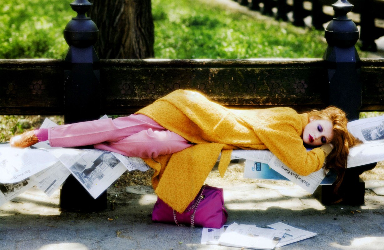 A woman in pink pants and a yellow coat rests on a park bench covered in newspapers next to a pink purse in a public park.