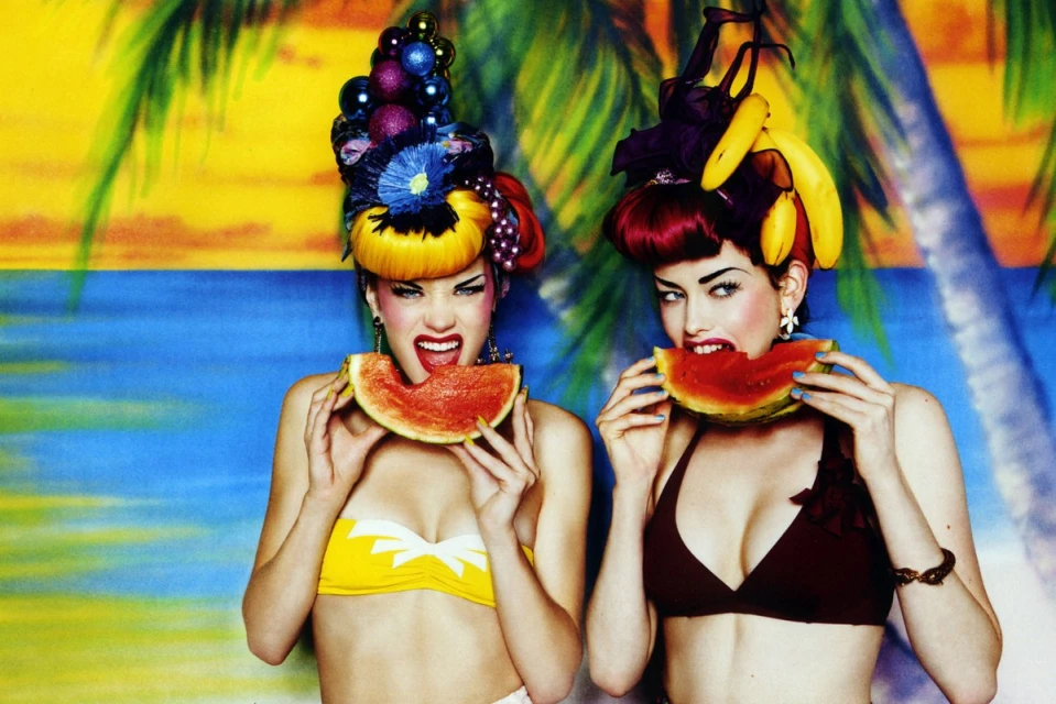 Two women in bikinis hold watermelon slices against a tropical backdrop, featuring fruit-themed hairstyles and bright summer colours.