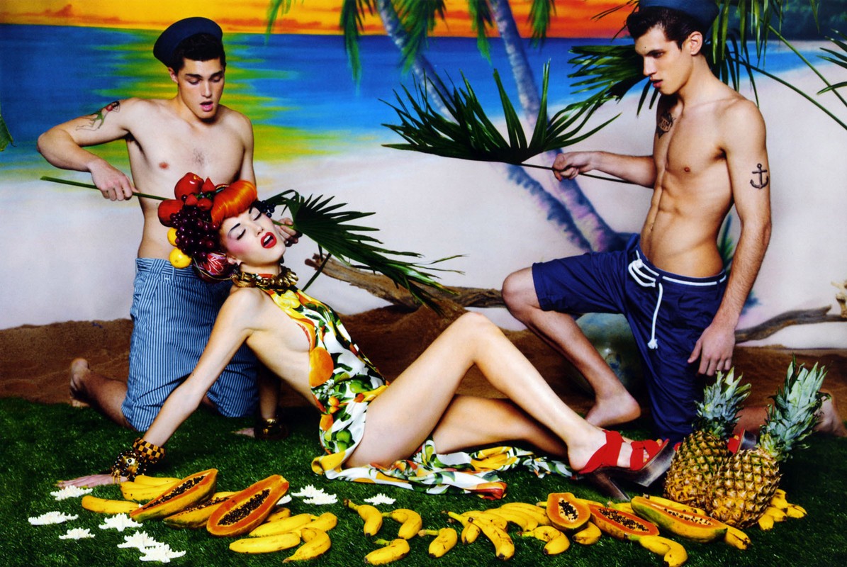 Eccentric fashion shoot featuring a woman with a fruit headdress, two male models, tropical fruit, and a beach backdrop.