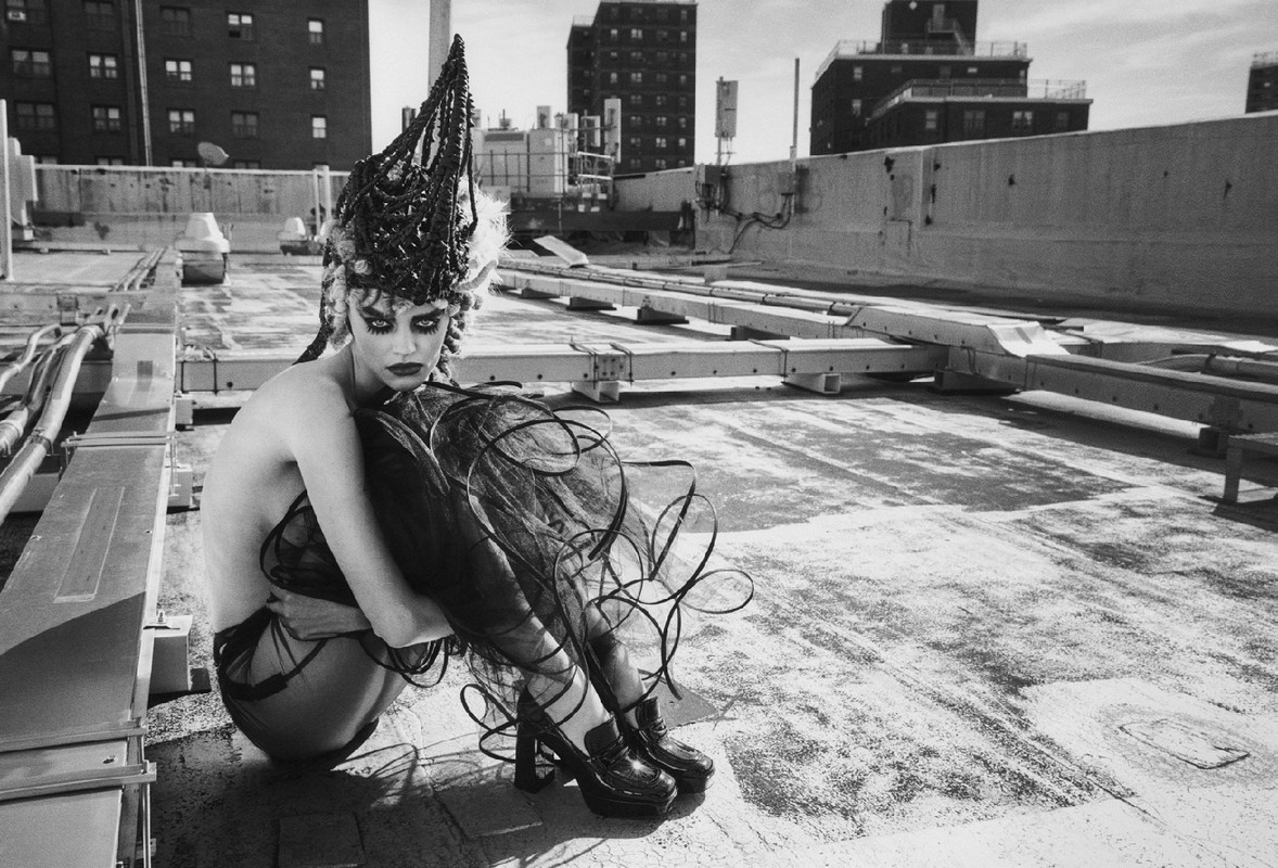 Dramatic monochrome fashion portrait on a rooftop features a model in avant-garde headwear and platform shoes against a cityscape backdrop.