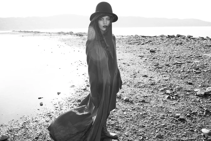 Monochrome portrait of woman in cloak and hat standing on rocky beach, gazing at camera, with water and distant mountains.